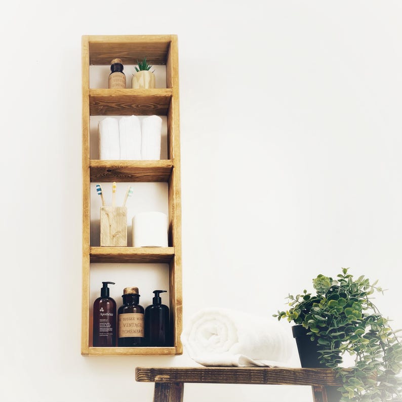 May include: A wooden bathroom shelf with four shelves. The top shelf has a small bottle and a potted plant. The second shelf has two white towels. The third shelf has a small wooden box with toothbrushes and a white candle. The bottom shelf has three bottles of liquid soap. A white towel is on the counter below the shelf.