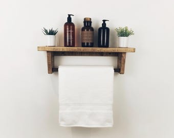 May include: A rustic wooden shelf with a towel bar, holding a white towel. The shelf displays two small potted plants and three bottles with pump dispensers. The shelf is mounted on a white wall.