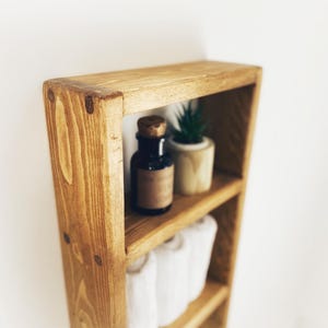 May include: A wooden shelf unit with three shelves. The top shelf has a small potted succulent plant and a dark glass bottle with a cork stopper. The middle shelf has white towels rolled up. The shelf is made of natural wood with a rustic finish.
