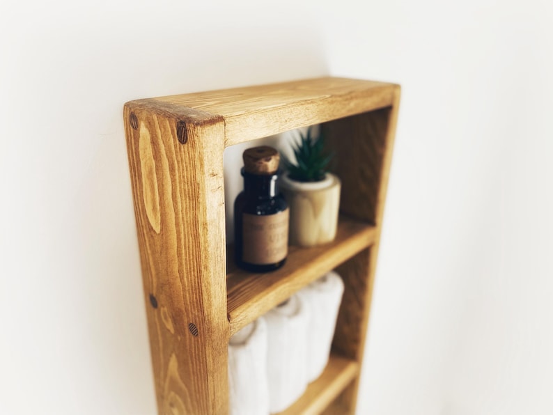 May include: A wooden shelf unit with three shelves. The top shelf has a small potted succulent plant and a dark glass bottle with a cork stopper. The middle shelf has white towels rolled up. The shelf is made of natural wood with a rustic finish.