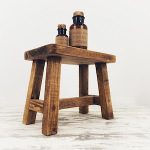 May include: A rustic wooden stool with a brown finish. Two small glass bottles with labels are sitting on top of the stool. The label on the larger bottle reads "Vintage Homeware".