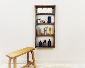 May include: A rustic wooden shelf unit with four shelves, featuring various bathroom essentials, including toiletries, hand towels, and decorative items. The shelf is mounted on a white wall.