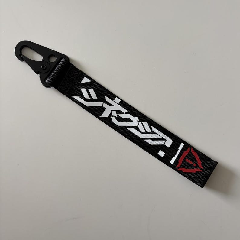 May include: Black keychain strap with white Japanese text and a red triangle logo. The strap has a black metal clip for attaching to keys or other items. The text is stylized and modern.