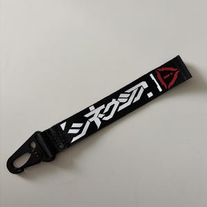 May include: Black lanyard with white Japanese text and a red graphic. The lanyard has a black metal clip. The text is in a stylized font. The lanyard is on a white surface.
