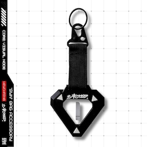 May include: Black keychain with a diamond-shaped pendant and a metal clasp. The pendant has Japanese text and white triangular accents. A black strap connects the pendant to the clasp and keyring. The text "CORE: VISUAL NODE" and "SYN. BAG ACCESSORY" are on the side.
