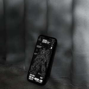 May include: Black phone case featuring a detailed white schematic of a robot, with text including "RX-93-2 HI-V" and "SERIAL RX932-EX/ ALPHA-VX". The case is angled against a textured gray surface.