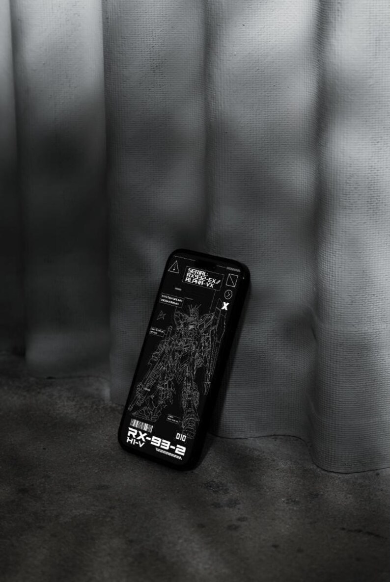 May include: Black phone case featuring a detailed white schematic of a robot, with text including "RX-93-2 HI-V" and "SERIAL RX932-EX/ ALPHA-VX". The case is angled against a textured gray surface.