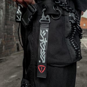 May include: Black tactical cargo pants with a strap featuring white Japanese text and a red triangle symbol. The pants have multiple pockets, a black chain, and a metal clasp.