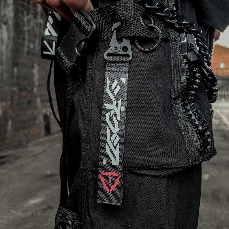 May include: Black tactical cargo pants with a strap featuring white Japanese text and a red triangle symbol. The pants have multiple pockets, a black chain, and a metal clasp.