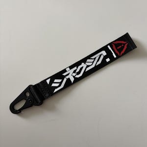 May include: Black lanyard with white Japanese characters and a red triangle design. The lanyard has a black metal clasp for attaching to keys or other items. The text is clear and easy to read.