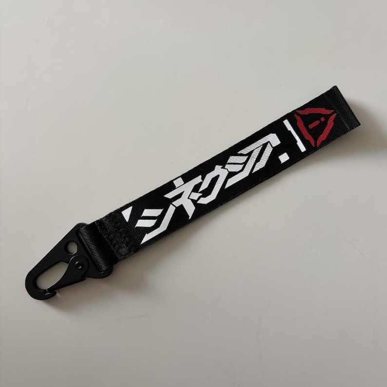 May include: Black lanyard with white Japanese characters and a red triangle design. The lanyard has a black metal clasp for attaching to keys or other items. The text is clear and easy to read.