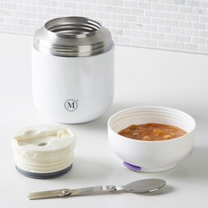 May include: White stainless steel food flask with a lid and a separate white bowl with a purple lid. The bowl contains a red soup with vegetables. A silver spoon is also included.
