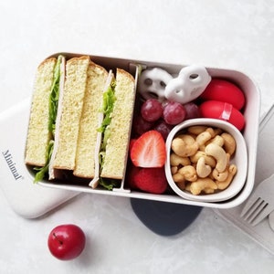 May include: A rectangular beige bento box with the word Minimal printed on the lid. The container holds sliced sandwiches with lettuce, red grapes, a halved strawberry, white yogurt-covered pretzels, two red round snacks, and a small bowl of cashews.