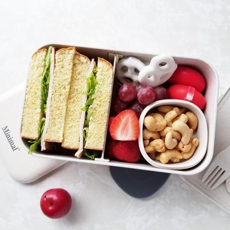 May include: A rectangular beige bento box with the word Minimal printed on the lid. The container holds sliced sandwiches with lettuce, red grapes, a halved strawberry, white yogurt-covered pretzels, two red round snacks, and a small bowl of cashews.