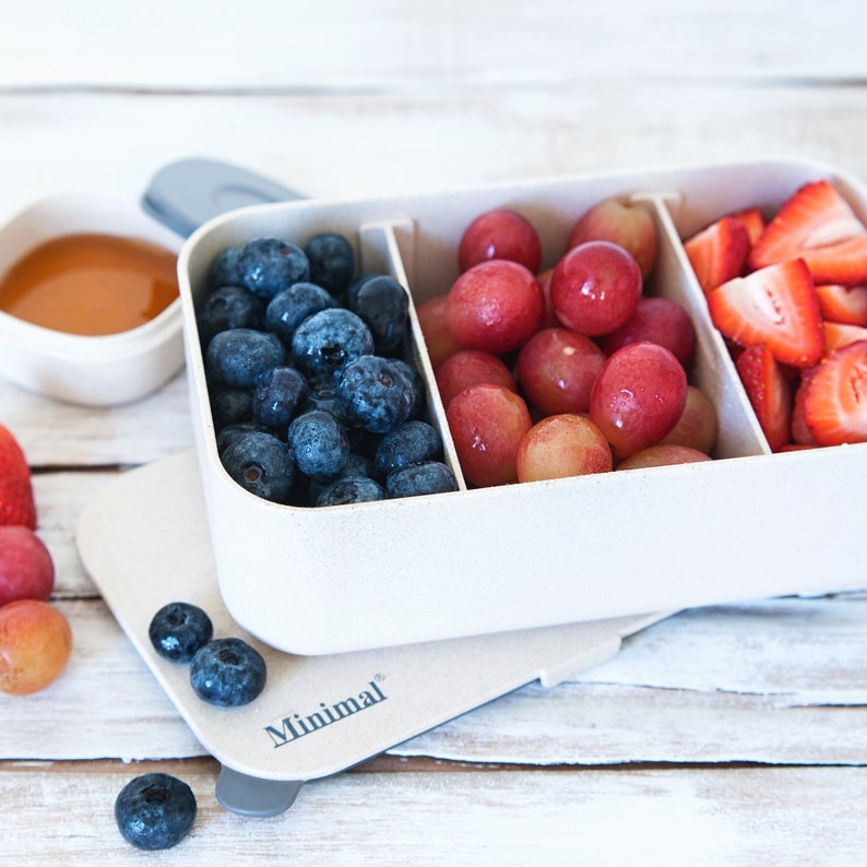 May include: A rectangular, off-white, three-compartment food container with the word Minimal printed on the lid. The compartments are filled with fresh blueberries, red grapes, and sliced strawberries. A small separate bowl contains honey.