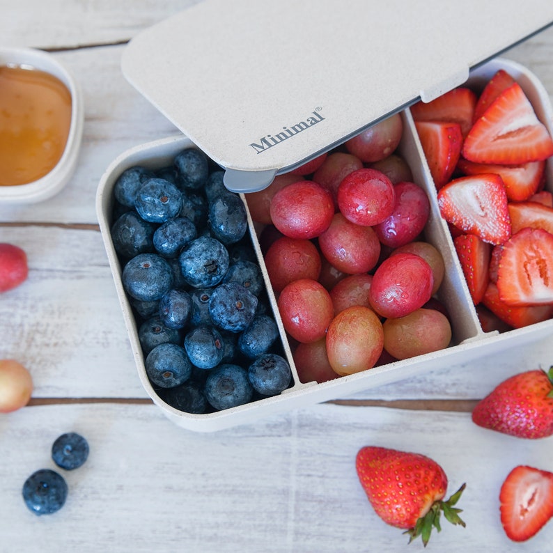 May include: A rectangular beige bento box with the word Minimal printed on the lid. The container is divided into two sections, one filled with fresh blueberries and the other with red grapes and sliced strawberries, with a small side dish of dip.