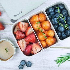 May include: A rectangular light gray bento box with the word Minimal printed on the lid, divided into three sections containing sliced strawberries, melon balls, and blueberries, with a small separate container of light brown sauce nearby.