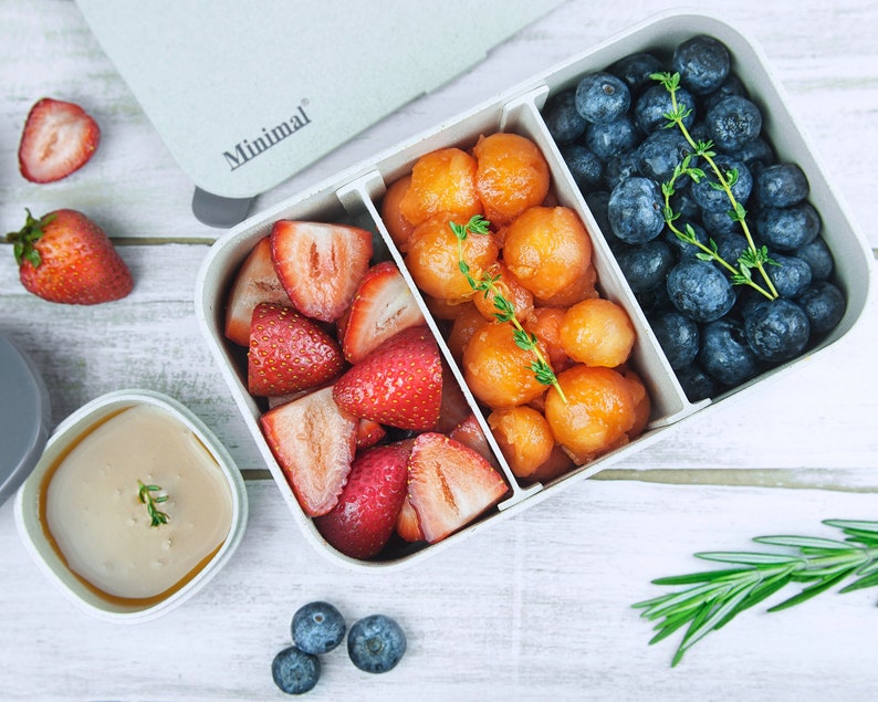 May include: A rectangular light gray bento box with the word Minimal printed on the lid, divided into three sections containing sliced strawberries, melon balls, and blueberries, with a small separate container of light brown sauce nearby.