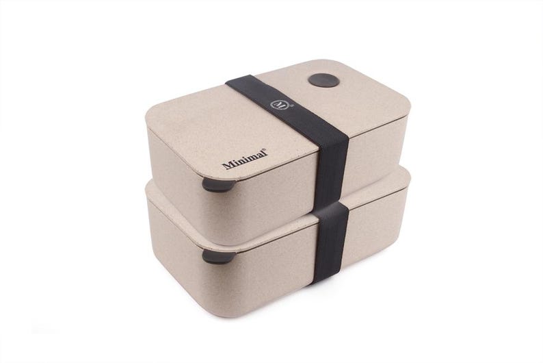 May include: A stack of two rectangular beige bento boxes with rounded corners. A black elastic band secures the containers together. The top lid features a circular brown accent and the text Minimal with a logo on the band and the side.