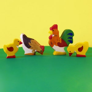 Op de afbeelding: Een set houten boerderijdieren speelgoed, inclusief een haan, een hen en twee kuikens. De haan is rood, geel en groen, de hen is bruin en wit, en de kuikens zijn geel. Het speelgoed is allemaal naar voren gericht en is in een rij op een groene ondergrond gerangschikt.