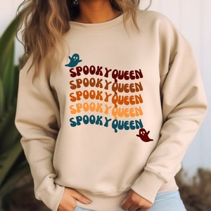 Può includere: Una felpa beige con il testo "Spooky Queen" ripetuto cinque volte in un arcobaleno di colori. Il testo è in un font retrò ed è accompagnato da una grafica di un fantasma.