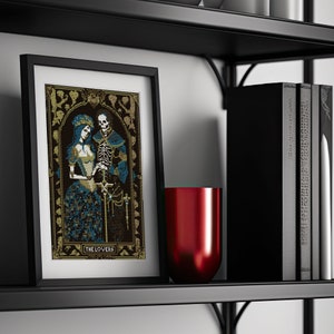 A digital render of 'The Lovers' Tarot cross stitch design. Presented in the format of a framed and mounted picture on a wooden shelf with vblack books and a metallic red vase. Strong black, white and blue DMC thread colours.