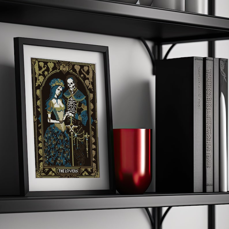 A digital render of 'The Lovers' Tarot cross stitch design. Presented in the format of a framed and mounted picture on a wooden shelf with vblack books and a metallic red vase. Strong black, white and blue DMC thread colours.