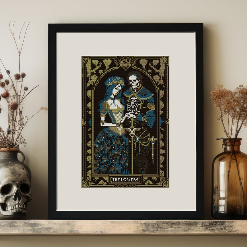 A digital render of 'The Lovers' Tarot cross stitch design. Presented in the format of a framed and mounted picture on a wooden shelf with vases, one shaped like a skull, and dried flowers. Strong black, white and blue DMC thread colours.