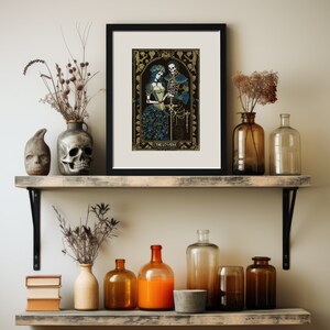 A digital render of 'The Lovers' Tarot cross stitch design. Presented in the format of a framed and mounted picture on a wooden shelf with vases, one shaped like a skull, and dried flowers. Strong black, white and blue DMC thread colours.