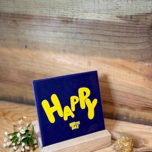 Könnte beinhalten: Eine quadratische, dunkelblaue Keramikfliese mit dem gelben Schriftzug "HAPPY" in einer großen, abgerundeten Schriftart und dem kleineren gelben Text "BIRTH DAY" darunter, auf einem hellen Holzständer mit kleinen Trockenblumen.