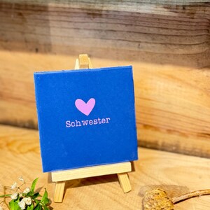 Könnte beinhalten: Eine quadratische blaue Leinwand auf einer kleinen Holzstaffelei. Die Leinwand zeigt ein hellrosa Herzsymbol über dem Wort "Schwester", das in einer passenden hellrosa serifenlosen Schrift mittig auf der blauen Fläche aufgedruckt ist.