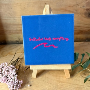 Könnte beinhalten: Eine blaue quadratische Leinwand mit dem Text "Saltwater heals everything" in Pink und einem pinkfarbenen Wellenmuster. Die Leinwand wird auf einer kleinen Holzstaffelei ausgestellt, mit Trockenblumen und Grünzeug im Hintergrund.