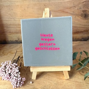 Könnte beinhalten: Ein graues quadratisches Schild mit den Worten "Heute wegen gestern geschlossen" in rosa Schrift, ausgestellt auf einer kleinen Holzstaffelei. Das Schild steht vor einem Holzgrund, mit rosa Blumen und grünen Blättern.