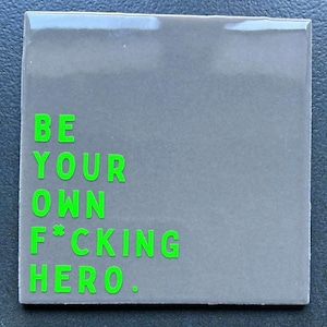 Könnte beinhalten: Quadratische, graue Fliese mit dem Satz "BE YOUR OWN F*CKING HERO." in leuchtend grünen, fetten Buchstaben. Die Fliese hat eine glänzende Oberfläche und ein schlichtes, modernes Design. Der Text ist auf der Fliese zentriert.