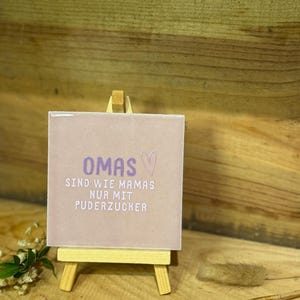 Könnte beinhalten: Eine quadratische, hellviolette Keramikfliese auf einer kleinen Holzstaffelei. Die Fliese zeigt den violetten Schriftzug OMAS, ein Herzsymbol und den weißen Text SIND WIE MAMAS NUR MIT PUDERZUCKER, was bedeutet, dass Omas wie Mamas sind, nur eben mit Puderzucker.