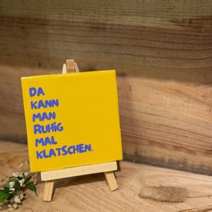 Könnte beinhalten: Ein quadratisches gelbes Schild mit blauem Text, der DA KANN MAN RUHIG MAL KLATSCHEN lautet. Das Schild steht auf einer kleinen hölzernen Staffelei. Die Oberfläche des Schildes ist glatt und glänzend und bildet einen Kontrast zum Holzständer.