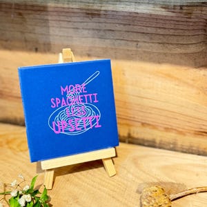 Könnte beinhalten: Ein quadratisches blaues Schild auf einer kleinen Holzstaffelei. Das Schild zeigt eine weiße Strichzeichnung einer Gabel, die Spaghetti aufdreht, und einen rosa Text mit der Aufschrift MORE SPAGHETTI LESS UPSETTI. Das Design ist mittig platziert.