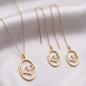 May include: A gold-colored jewelry set featuring a necklace and a pair of earrings. The necklace has a pendant with a crown design inside an oval frame. The earrings are threader style, with matching crown pendants. The set is displayed on a white satin surface.