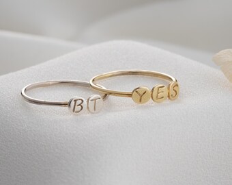 Op de afbeelding: Twee fijne ringen, één zilverkleurig en één goudkleurig, worden getoond. De zilveren ring spelt "BT", terwijl de gouden ring "YES" spelt. De ringen hebben dunne banden met kleine, cirkelvormige letters.