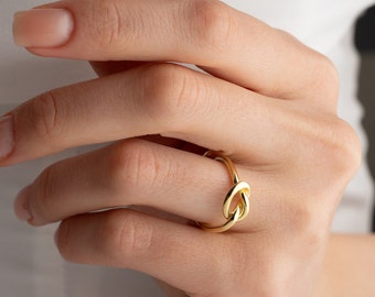 May include: A gold knot ring with a simple design. The ring is worn on a finger.