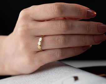 Op de afbeelding: Een goudkleurige ring met de letters "YES" gegraveerd op drie kleine, ronde bedels. De ring is dun en delicaat en wordt aan een vinger gedragen. De achtergrond is wazig, met een hand en een boek zichtbaar.