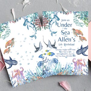 Pu&ograve; includere: Invito di compleanno a tema mare con un design ad acquerello blu e bianco che presenta una variet&agrave; di creature marine, tra cui uno squalo, un polpo, una tartaruga e una razza. L'invito include il testo "Unisciti a noi sotto il mare per il 5&deg; compleanno di Allen!" e dettagli sulla festa, tra cui la data, l'ora e la posizione.