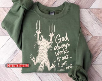 Może przedstawiać: Oliwkowa bluza z okrągłym dekoltem z białą grafiką kota wiszącego na pazurach. Tekst brzmi: God always Works it out... I just panic first. Projekt zawiera ślady pazurów nad kotem oraz tekst umieszczony po prawej stronie grafiki.