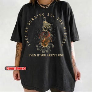 May include: A black short-sleeve t-shirt featuring a gold graphic of a figure standing above a fire with logs. The text reads "THEY'RE BURNING ALL THE WITCHES" in an arc above and "EVEN IF YOU AREN'T ONE" below the central illustration.