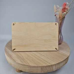 May include: A blank wooden sign with gold hanging hardware sits on a wooden pedestal. The sign is rectangular and has a light wood grain. The pedestal is round and has a natural wood finish.
