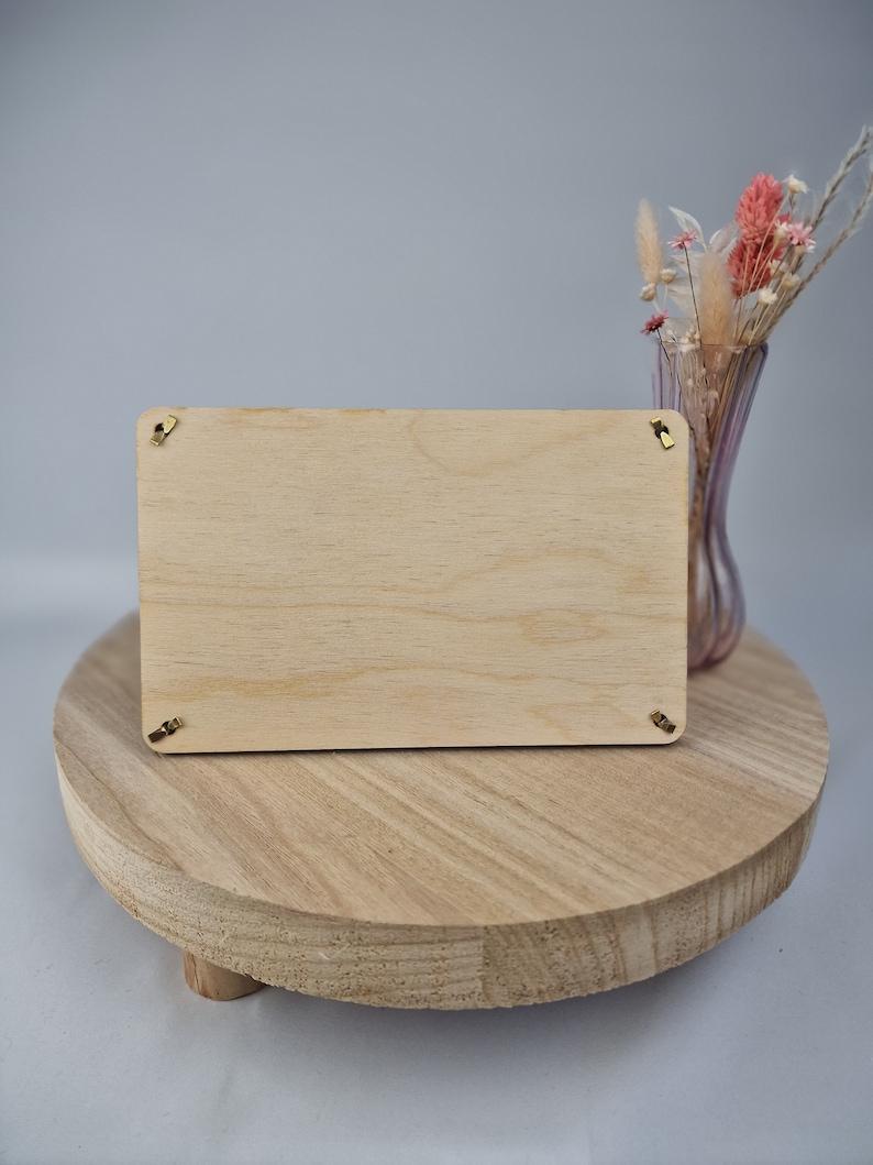 May include: A blank wooden sign with gold hanging hardware sits on a wooden pedestal. The sign is rectangular and has a light wood grain. The pedestal is round and has a natural wood finish.