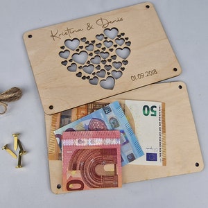 May include: A wooden box with a heart-shaped cutout and the text "Kristina & Denis 01.09.2018" engraved on the top. The box is open and there are several euro bills inside.