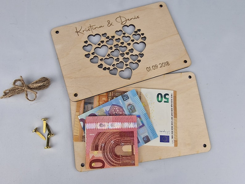 May include: A wooden box with a heart-shaped cutout and the text "Kristina & Denis 01.09.2018" engraved on the top. The box is open and there are several euro bills inside.