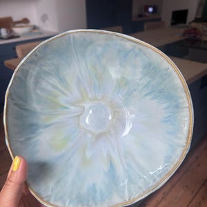 May include: A round ceramic bowl featuring a glossy glaze with swirling patterns of light blue, white, and pale yellow. The bowl has an irregular, organic rim finished with a thin, brownish-gold edge that contrasts with the cool-toned interior.