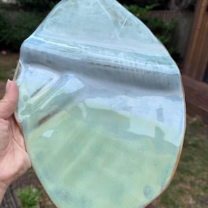 May include: An irregular, organic-shaped ceramic plate featuring a glossy glaze in shades of pale mint green and soft blue. The surface has a smooth, reflective finish with subtle colour variations and a visible brown clay rim along the edge.